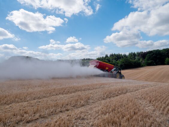 En traktor sprer kalk på en høstet kornåker under en blå himmel med skyer.