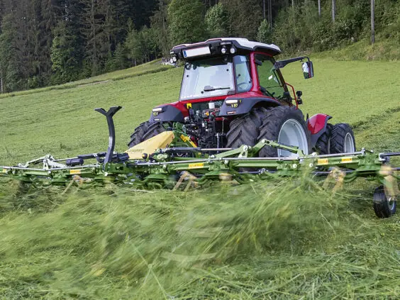 Rød traktor i arbeid på en gressbakke med en KRONE Vendro Highland høytørker.