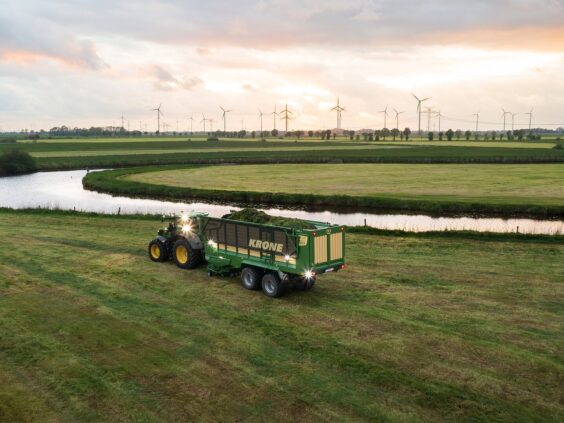 En traktor trekker en full KRONE ZX lessevogn på et grønt jorde ved solnedgang.