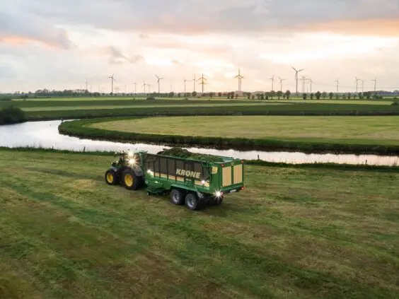 En traktor trekker en full KRONE ZX lessevogn på et grønt jorde ved solnedgang.