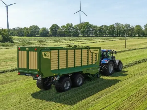 En blå traktor som trekker en grønn KRONE RX lessevogn på et jorde.