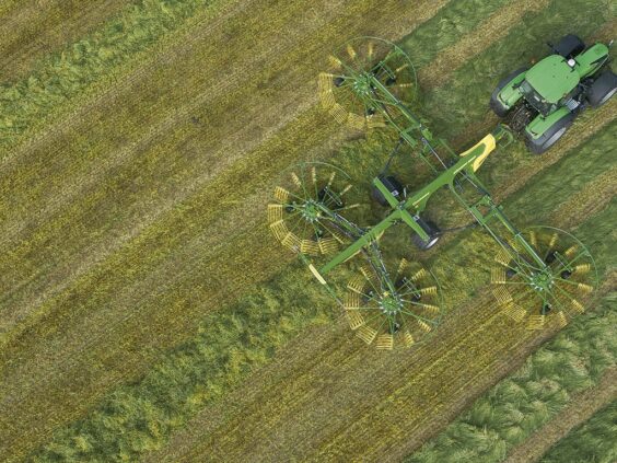 En grønn traktor trekker en KRONE Swadro TC samlerive på et jorde sett ovenfra.