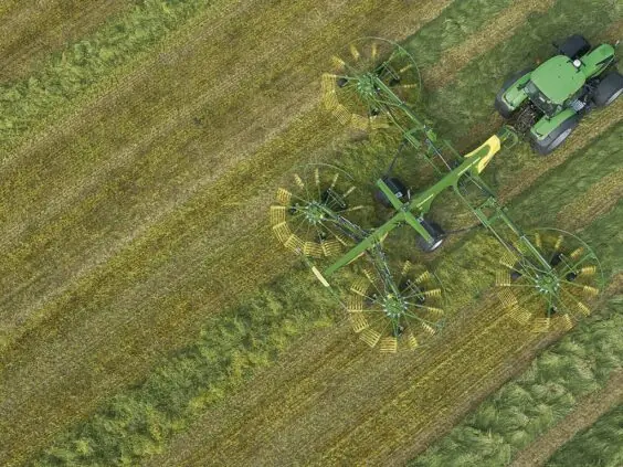 En grønn traktor trekker en KRONE Swadro TC samlerive på et jorde sett ovenfra.