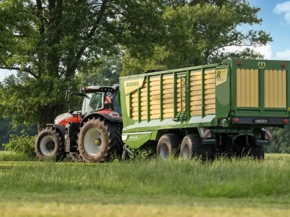 En rød traktor trekker en grønn og gul KRONE RX lessevogn på et jorde.