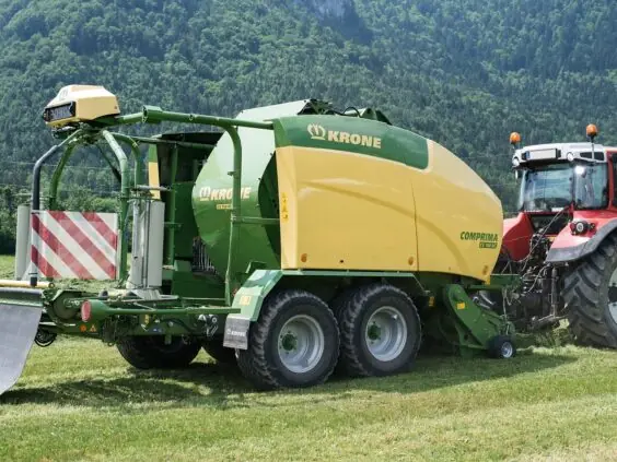 En rød traktor som trekker en Krone Comprima rundballepresser på et grønt jorde.