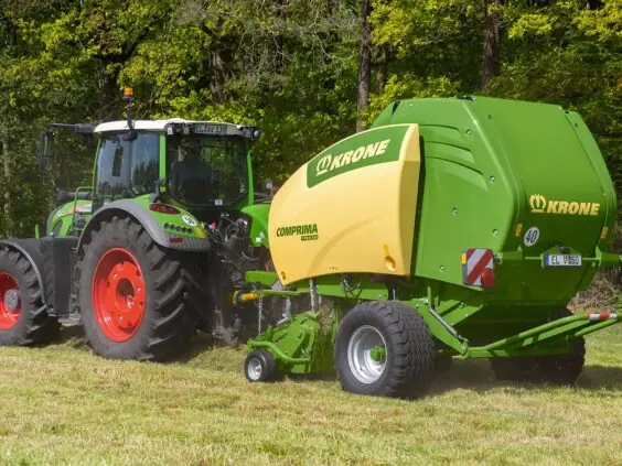 En traktor som trekker en grønn og gul Krone Comprima rundballepresser på et gressfelt.