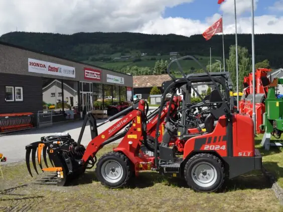 Rød Schäffer minilaster utstilt utenfor Fron Traktor Service sine lokaler i naturskjønne omgivelser.