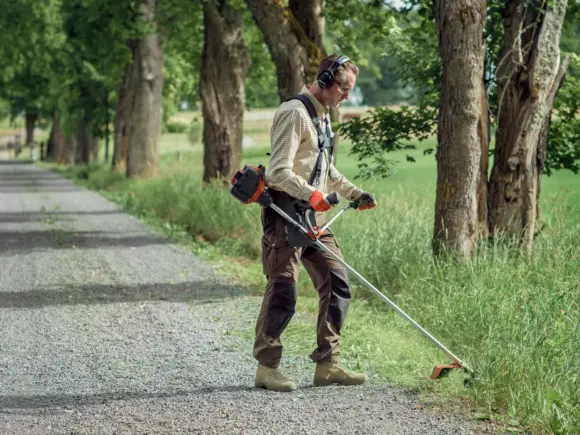 En mann bruker en gresstrimmer til å klippe høyt gress langs en grusvei.