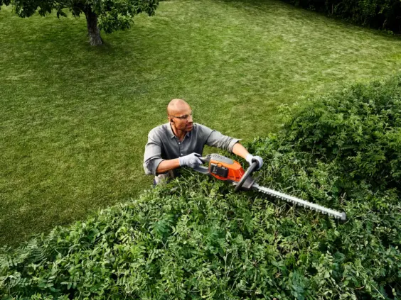 En mann som trimmer en grønn hekk med en batteridrevet hekksaks.