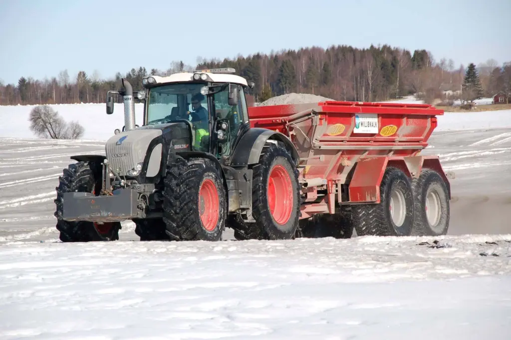 En grå traktor med rød Bredal kalkspreder kjører på et snødekket jorde om vinteren.