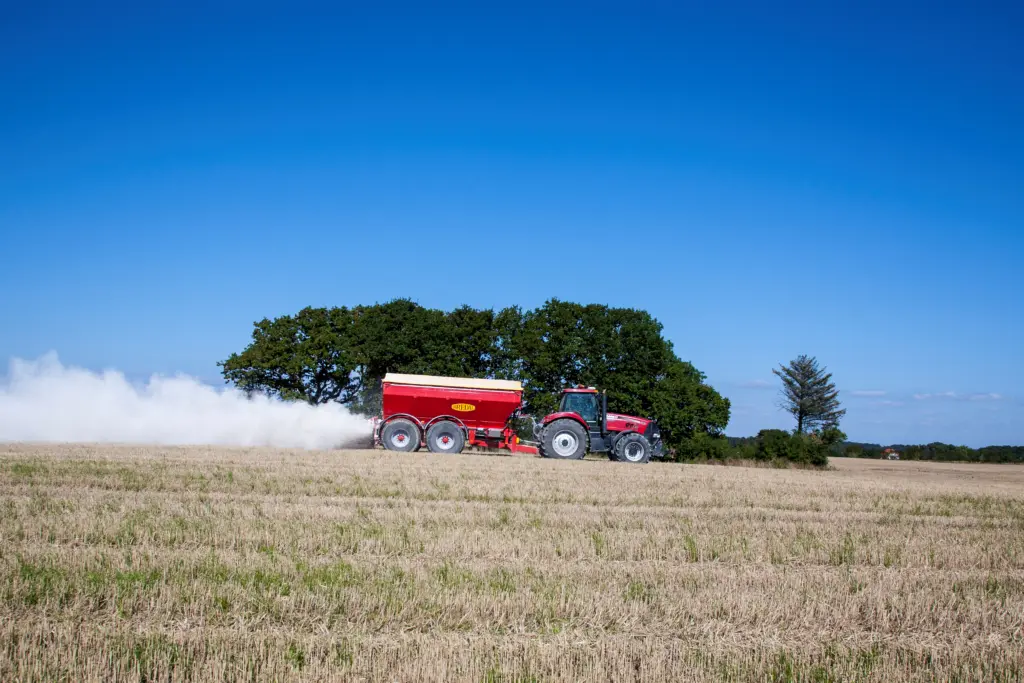 Rød traktor som sprer kalk på en åker under en klar blå himmel.