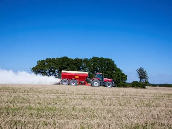 Rød traktor som sprer kalk på en åker under en klar blå himmel.
