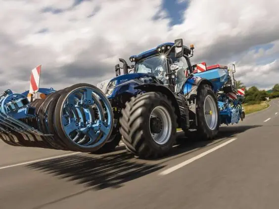 Ny New Holland T7 Standard Wheelbase-traktor på asfaltert vei med landbruksutstyr.