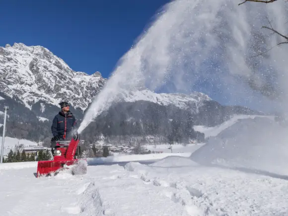 Rød snøfreser som kaster snø høyt i luften foran snødekte fjell.