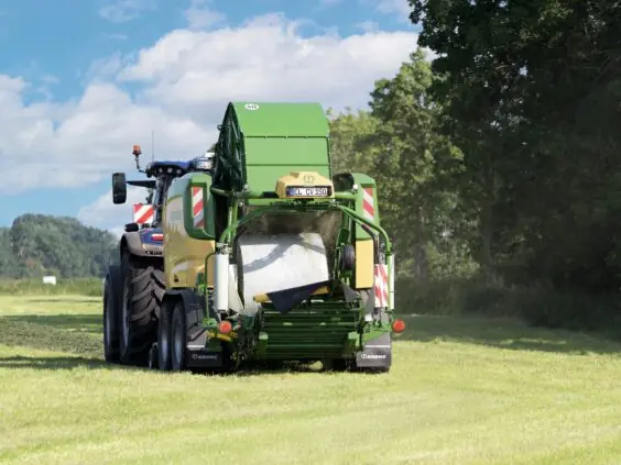 KRONE ComPack CV 150 XC Pro pakker en rundball i folie bak en blå New Holland-traktor på nyslått eng
