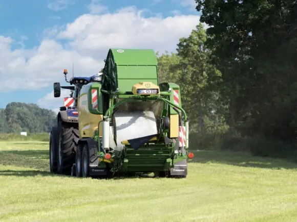 KRONE ComPack CV 150 XC Pro pakker en rundball i folie bak en blå New Holland-traktor på nyslått eng